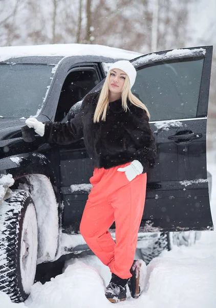 На відкритому повітрі зимовий портрет усміхненої блондинки біля чорної машини. Сніг навколо . — стокове фото
