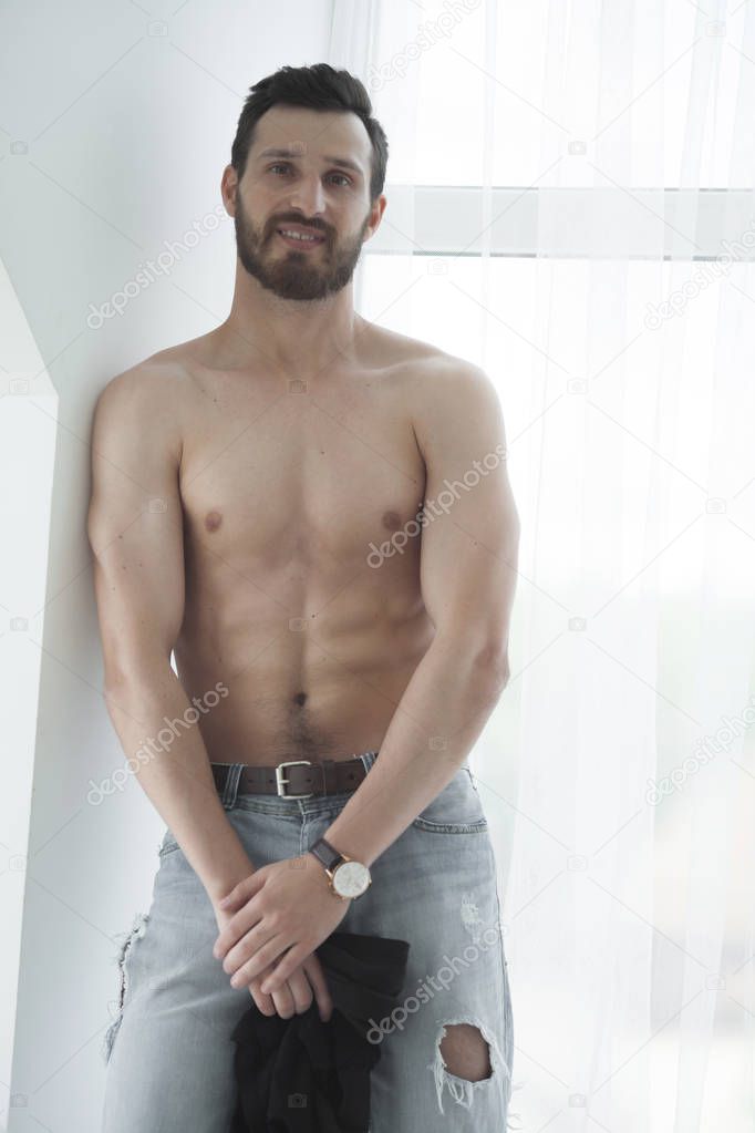 Sexy fashion portrait of a hot bearded male model in stylish jeans with muscular body posing in modern interior setting with window light