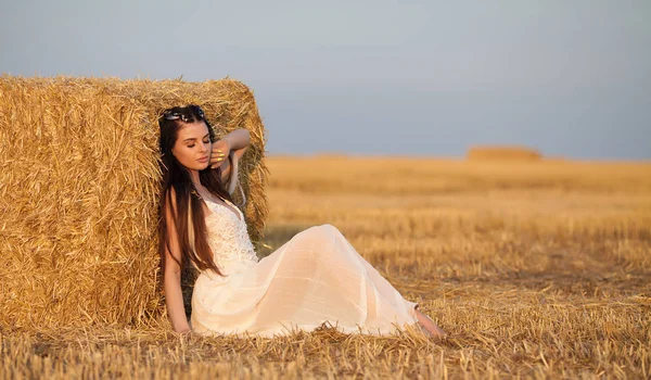 attractive stylish Woman in long white summer dress near hay bale on meadow