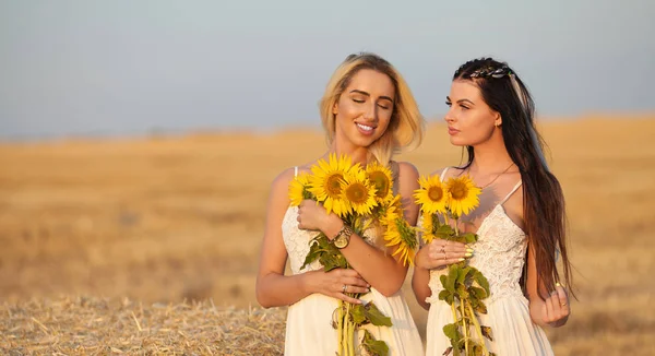 Två flickvänner blondin och brunett i fältet med solrosor i hand. — Stockfoto