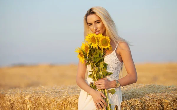 Mooie lieve sexy meid in een witte jurk lopen op een veld zonnebloemen in de hand. — Stockfoto