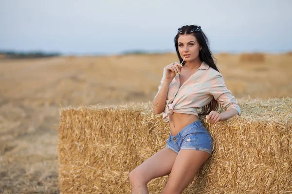Sexy brunette woman in a bra and jeans shorts posing and smiling against a stack of hay in a summer day — Stock Photo, Image