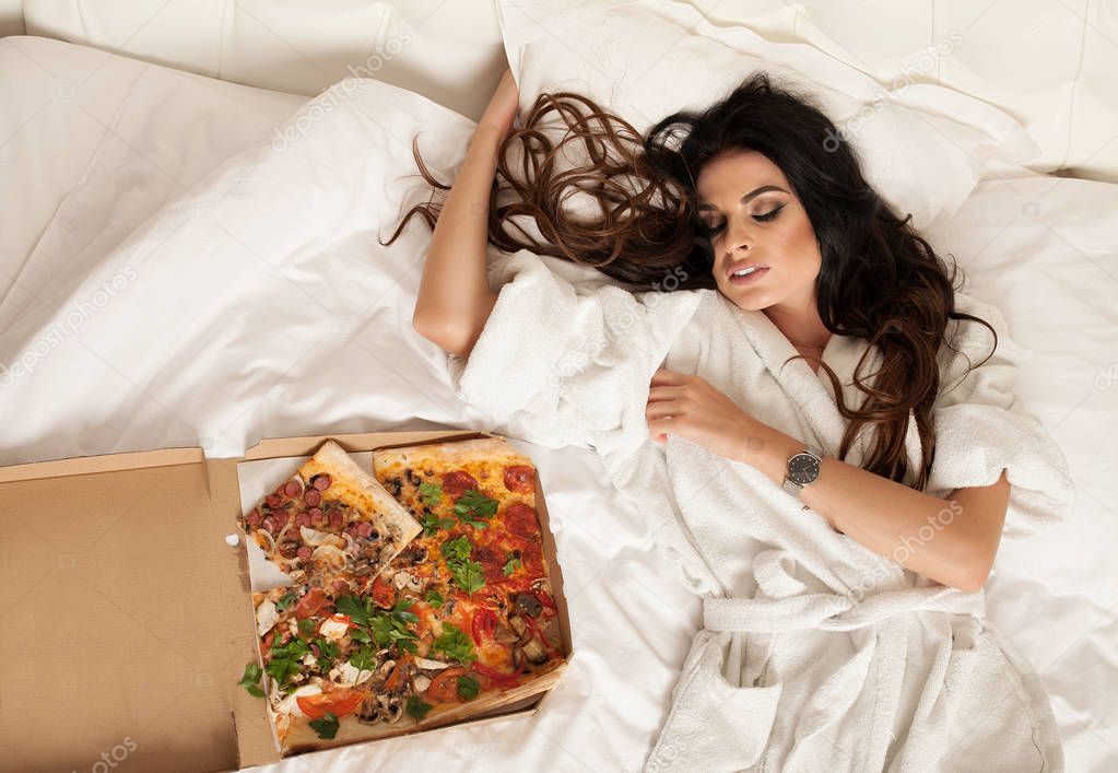 Pretty young sexy fashion sensual woman posing on white background.Sensual woman in white bathrobe. White bed isolated, crazy night party time,girl eating tasty pizza