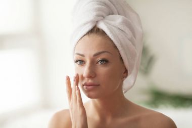 Genç kadın kusursuz bir cilt, yüzünde nemlendirici krem uygulamak. Kadın beyaz bornoz ve havlu banyoda sonra fotoğrafı. Cilt bakım kavramı