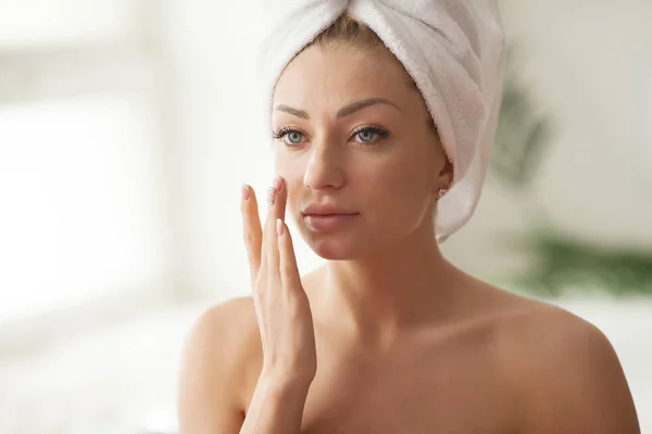 Young woman with flawless skin, applying moisturizing cream on her face. Photo of woman after bath in white bathrobe and towel. Skin care concept