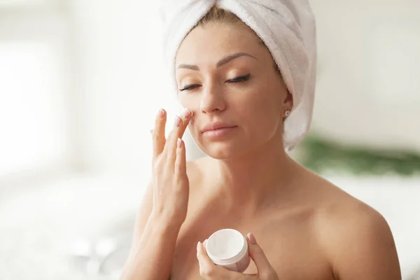 Young woman with flawless skin, applying moisturizing cream on her face. Photo of woman after bath in white bathrobe and towel. Skin care concept
