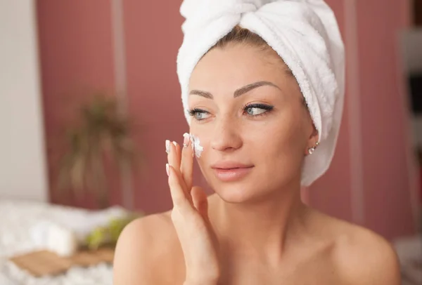 Young woman with flawless skin, applying moisturizing cream on her face. Photo of woman after bath in white bathrobe and towel. Skin care concept