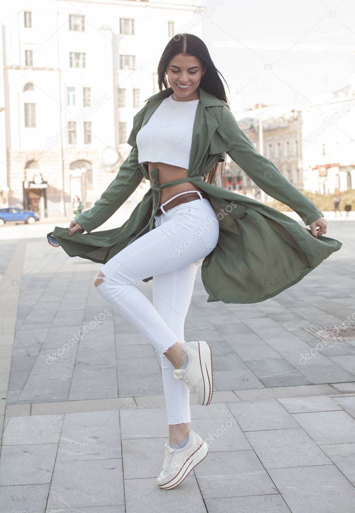 Beautiful young stylish girl in a fashion green coat, white pants and white sneakers in the street on a sunny day
