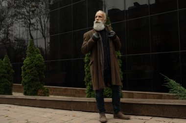 Sakallı yakışıklı, kendine güvenen olgun bir adam mesaj ya da reklam için fotokopi yeri olan paltoda duruyor. Pahalı kıyafetler giymiş, dışarıda poz veren şık zengin bir erkek..