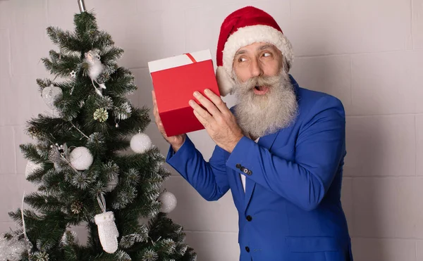 白い背景の上にギフトボックスを持っているハンサムなシニア髭の男とカメラを見て サンタクロースはメリークリスマスと幸せな新年を願っています 青いスーツの男クリスマス — ストック写真