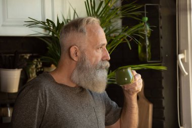 Yakışıklı yaşlı sakallı adam mutfakta kahve içiyor.