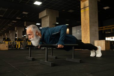 Beyaz sakallı, yakışıklı, kendini beğenmiş, beyaz, yaşlı bir adam göbek ve kollarını güçlendirmek için şınav çekiyor. Tahtada duran çekici, konsantre, emekli bir erkek. Fitness, spor, hobi ve etkinlik.