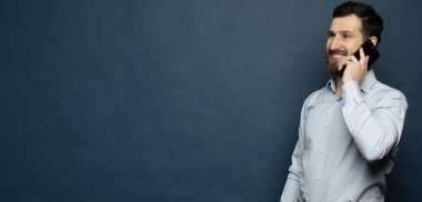 Mutlu gülümseyen genç adam mavi arka planda cep telefonuyla konuşuyor.