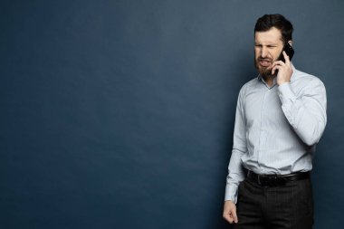 Sakallı bir adam telefonda bağırıyor ve konuşuyor. İzole edilmiş mavi arkaplan.