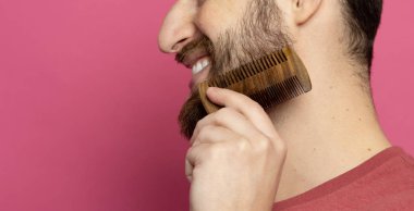 Young bearded man comb his beard and moustache on pink background