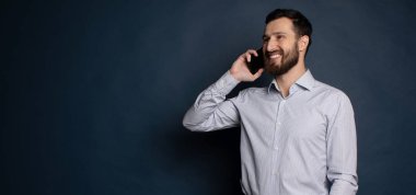 Mutlu gülümseyen genç adam mavi arka planda cep telefonuyla konuşuyor.