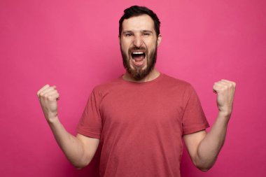 Young bearded man celebrating victory over pink background.