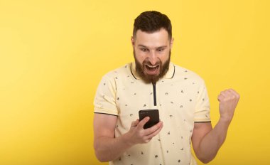 Portrait of a confident happy man holding mobile phone and celebrating success isolated over yellow background.