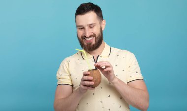 Mavi arka planda duran ve hindistan cevizi suyu içen çekici genç sakallı adamın portresi.