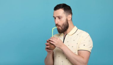 Mavi arka planda duran ve hindistan cevizi suyu içen çekici genç sakallı adamın portresi.