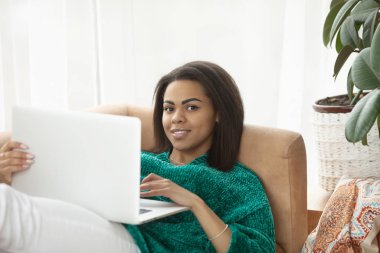Evde internette sörf yapmak. Genç Afrikalı kadın dizüstü bilgisayarda çalışıyor ve evinde kanepede otururken gülümsüyor.