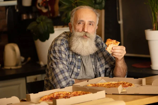 Smiling older man eating pizza slice sitting at living room. Bearded senior man eating italian food.
