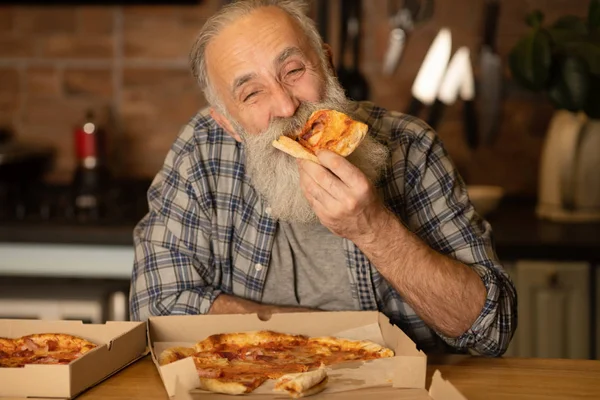 Smiling older man eating pizza slice sitting at living room. Bearded senior man eating italian food.