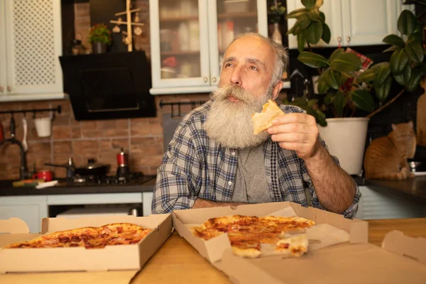 Smiling older man eating pizza slice sitting at living room. Bearded senior man eating italian food.