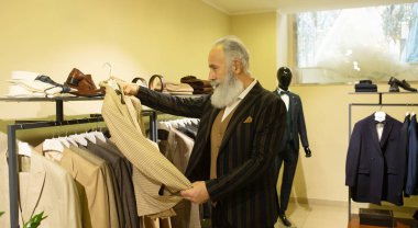 Giyim mağazasında kıyafet alışverişinde takım elbise seçen sakallı kıdemli bir adam..