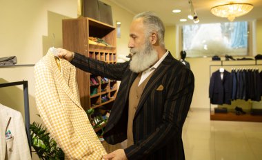 Giyim mağazasında kıyafet alışverişinde takım elbise seçen sakallı kıdemli bir adam..