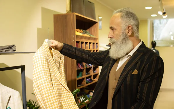 Bearded senior man choosing suit jacket during apparel shopping at clothing store.