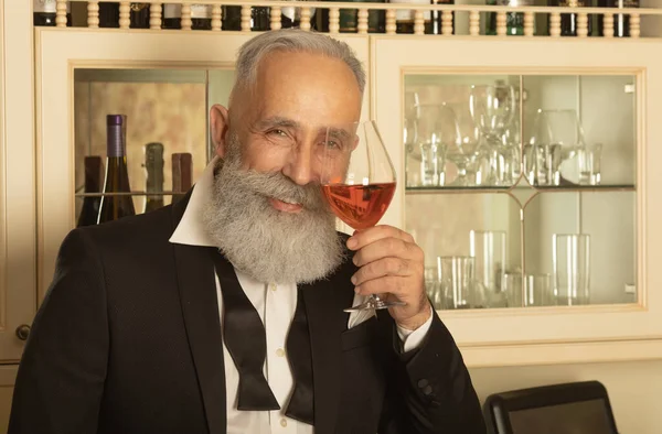 Elegant bearded senior gentleman proposing a toast with a glass of rose wine and looking at the camera.