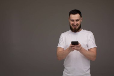 Yakışıklı sakallı model gri arka planda elinde telefonla poz veriyor..