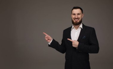 Genç başarılı esmer borsa komisyoncusunun yakın plan portresi gri arka planda, gülümsüyor, takım elbise giyiyor ve parmağıyla bir fotokopi alanını işaret ediyor..