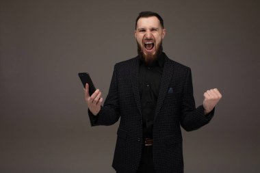 Mutlu işadamı akıllı telefon tutarak ve gri arka plan üzerinde başarısını kutluyor.