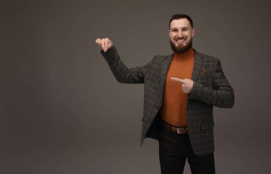 Genç başarılı esmer borsa komisyoncusunun yakın plan portresi gri arka planda, gülümsüyor, takım elbise giyiyor ve parmağıyla bir fotokopi alanını işaret ediyor..