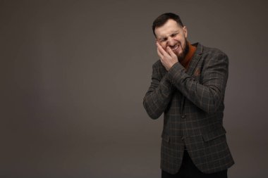 Sinirli, mutsuz, sorunlu, yakışıklı, sakallı adamın yanağına dokunan portresi. Gri arka planda diş ağrısı var..