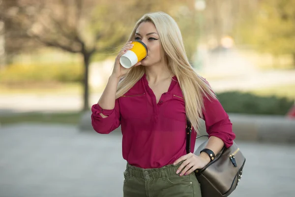 Cheerful fashionable blonde holding coffee outdoors.Young woman drinking coffee on the street of the city.