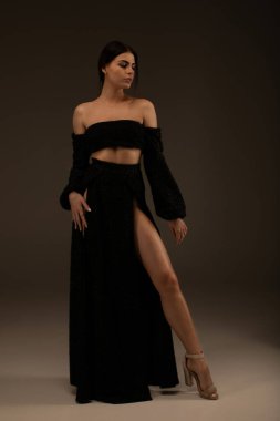 Gri arka planda stüdyoda poz veren uzun bacaklı, siyah giysili zarif güzel bir kadın..