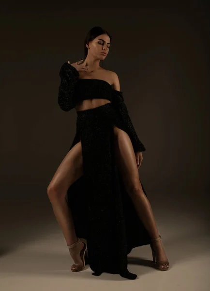Elegant beautiful woman with long legs in black fashionable dress posing in studio on grey background.