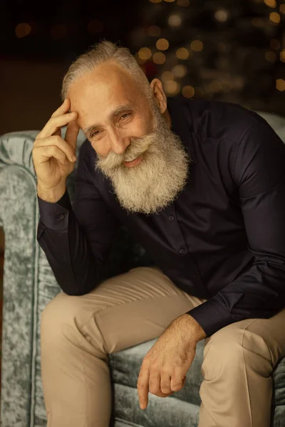 Positive elderly bearded gentleman smiling looking at camera sitting on sofa at home.