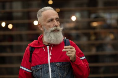 Sakallı yaşlı adam duman üflüyor. Elektronik sigara içiyorum. Stres atma konsepti. Sigara içme cihazı. Uzun sakallı adam sigara içmekten rahatlamış. Sakallı adam buharlaşıyor. IQOS.