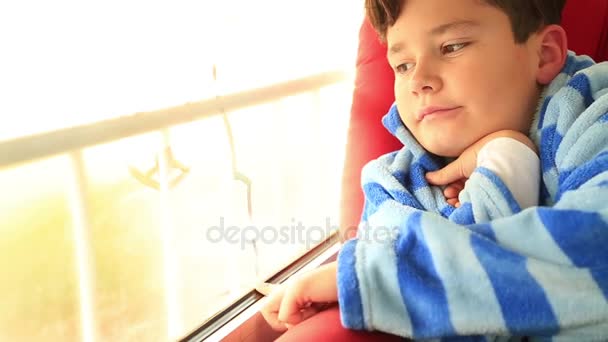 Een jonge jongen zit kijkt uit het raam — Stockvideo