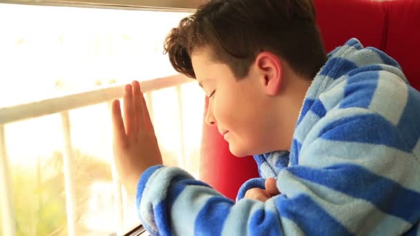 Ένα νεαρό αγόρι κάθεται κοιτάζοντας έξω από το παράθυρο — Αρχείο Βίντεο