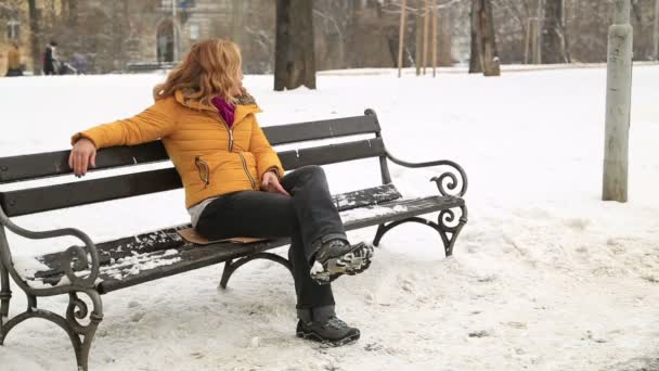 ベンチに座っていると 雪の降る屋外でカメラを見て満足し 笑みを浮かべて女性 — ストック動画