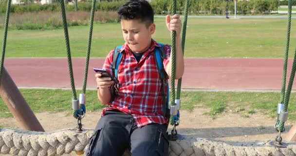 スマート フォンを使用して 遊び場でスイング幸せのプレティーンの少年の肖像画 の概念 スマート フォン中毒 インターネット通信技術 — ストック動画