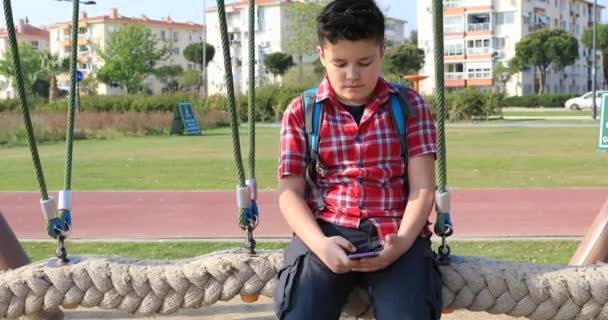 スマート フォンを使用して 遊び場でスイング幸せのプレティーンの少年の肖像画 の概念 スマート フォン中毒 インターネット通信技術 — ストック動画