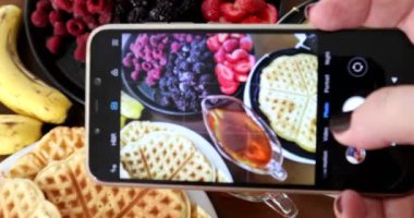 Kırmızı meyveli ve muzlu waffle 'ın fotoğraflarını çekmek için akıllı telefon kullanan kadın eller.