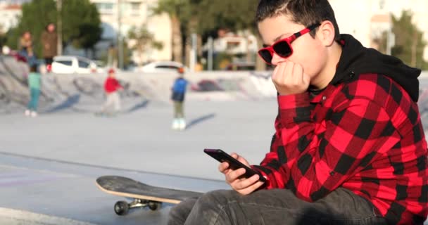 スケートパークの近くに座っているハンサムな10代の少年の肖像画だけで子供の画面を見て 読み取り ゲームをプレイ テクノロジー インターネット通信と人々の概念 スマートフォン中毒 — ストック動画