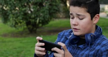 Portrait of a excited teen boy sitting on the park bench alone Kid watching to screen, reading, typing, playing games. Technology, internet communication and people concept, Smartphone addiction